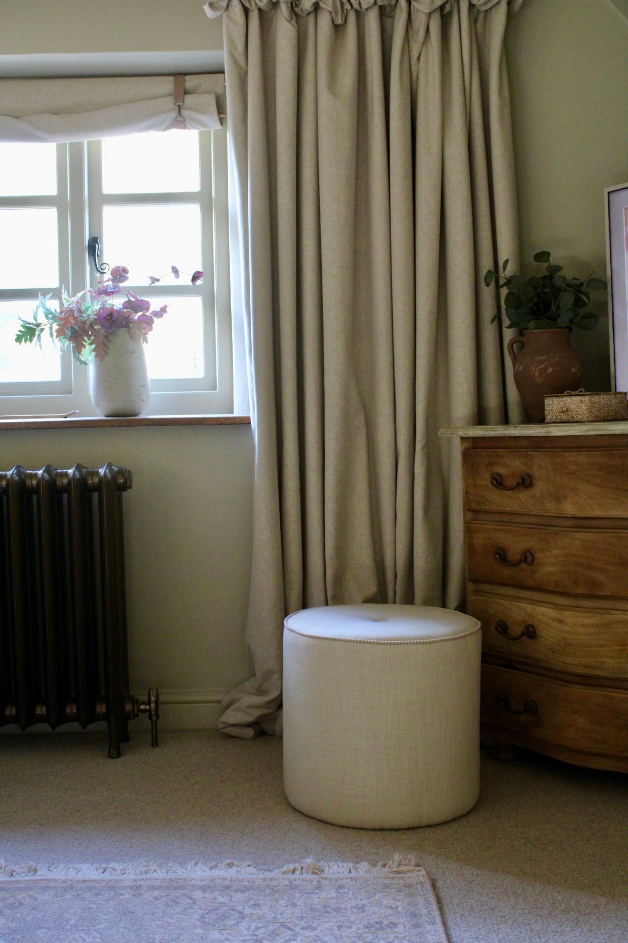 Room interior with a curtain, radiator, ottoman, and dresser.